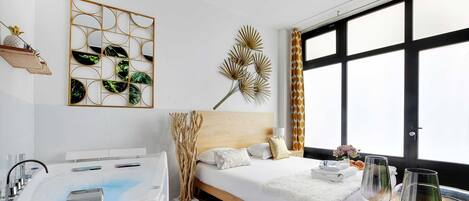 A modern hotel room with a large window, a bathtub, a bed with white linens, and decorative wall art.