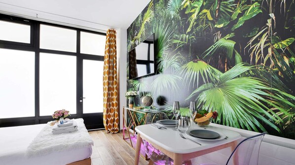 A modern room with a large window, a dining area with a tropical-themed mural, and a cozy seating area with a white rug and a small plant. - Magnificent Flat - 2br/6p - Champs Elysaes (Paris)