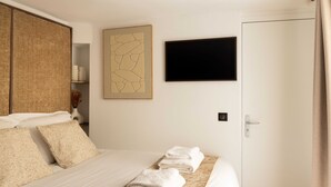 Een netjes opgemaakt bed met witte lakens en een opgevouwen handdoek, een wandtelevisie en een deur met gordijn.