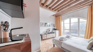 A modern bedroom with a wooden ceiling, a large window with curtains, a bed with neatly folded towels, and a kitchen area with a sink and utensils. - Apartment in Paris for 2 People (Paris)