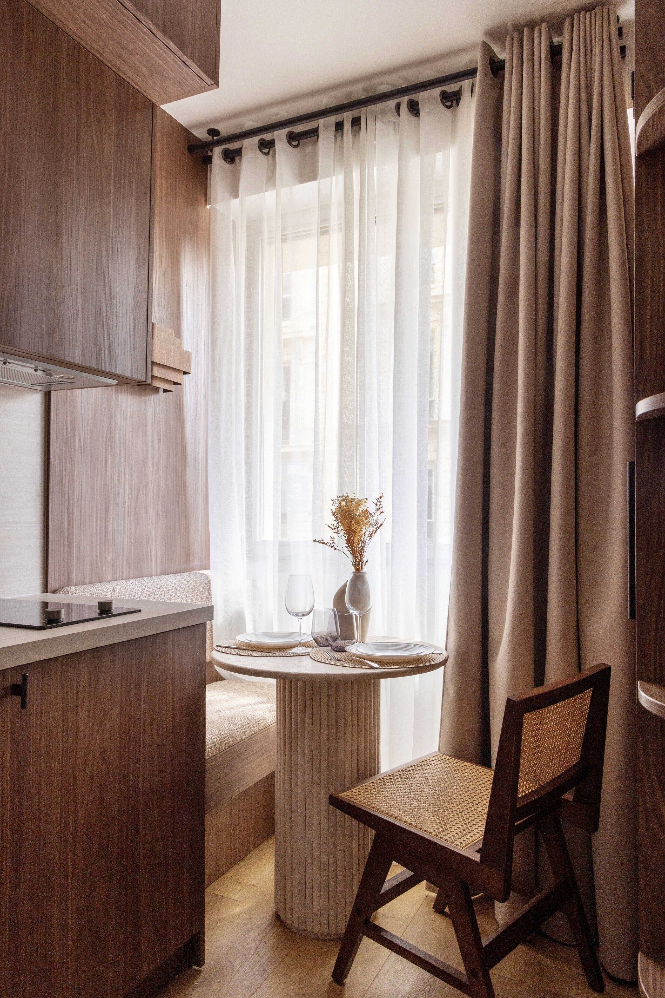 A compact kitchen with wooden cabinetry, a round dining table with a chair, and a window with sheer curtains.