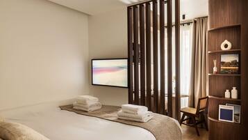 A hotel room with a bed, a desk, a chair, a TV, and a wooden shelf with books and decorative items.
