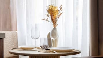 Un coin repas avec une table ronde dressée pour deux personnes, un vase contenant des fleurs séchées et une chaise dont le dossier est en osier tressé.