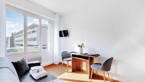A modern living room with a sofa, a dining table with chairs, a flat-screen TV, and a large window with a city view. - Charming Studio - 2P - Parc Georges Brassens (Paris)