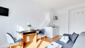 A modern living room with a grey sofa, a wooden dining table with two chairs, a kitchen area with a microwave, and a TV mounted on the wall. - Charming Studio - 2P - Parc Georges Brassens (Paris)