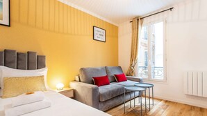 A hotel room with a bed, a grey sofa with red cushions, a small round table, and a window with a view of buildings. - Charming Studio - 4P - Place de la Bastille (Paris)