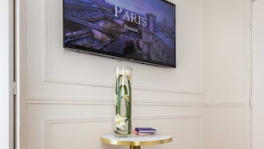 Ein modernes Zimmer mit einem runden goldenen Tisch, zwei goldenen Hocker, einem Wand-Fernseher, der eine Pariser Szene zeigt, und einer Vase mit Blumen.