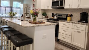 Une cuisine moderne avec des meubles blancs, une cuisinière en acier inoxydable et un four à micro-ondes. On y trouve un coin bar avec des tabourets, ainsi qu'un vase rempli de fleurs sur l'îlot central.