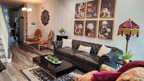 Un salon moderne avec un canapé en cuir noir, une table basse et un tapis à motifs. On y trouve deux fauteuils, une lampe sur pied et un élément décoratif fixé au mur. On aperçoit une cuisine au fond.