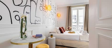Quarto de hotel com uma cama, uma mesa redonda com um vaso, um banquinho pequeno e uma parede com arte abstrata.
