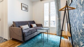 Una sala de estar con un sofá gris, dos mesitas redondas, una escalera de madera, una cortina estampada y un cuadro en la pared.