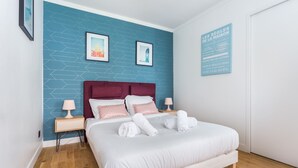 A neatly made bed with white linens and pink pillows, a wooden headboard, and a blue wall with geometric patterns. There are two framed pictures on the wall, a white door, and a sign with French text. - Peaceful Studio - 2P - Place de la Bastille (Paris)