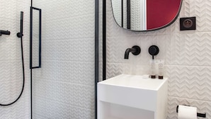 Un bagno moderno con box doccia in vetro, lavandino bianco con rubinetto nero e specchio con dettaglio rosso.