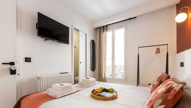 Una camera d'albergo con un letto, una TV a schermo piatto fissata al muro, un termosifone, una finestra con tende e una porta con maniglia.