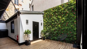 Un cortile con un pavimento in legno, un edificio bianco, una porta nera, un estintore, una pianta in vaso e un muro ricoperto di verde e fiori rossi.