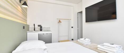 A modern hotel room with a bed, a TV mounted on the wall, a small kitchenette, and a bathroom visible through an open door.
