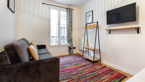 Una sala de estar con un sofá oscuro, alfombra colorida, un televisor montado en la pared y un ventanal con cortinas.