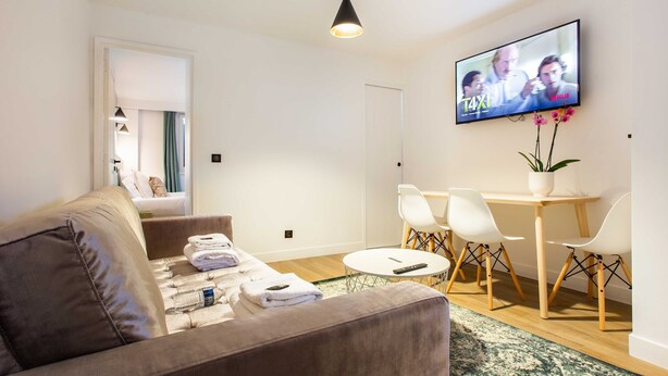 Ein modernes Wohnzimmer mit einer Couch, einem Esstisch mit Stühlen, einem an der Wand befestigten Fernseher und einer Zimmerpflanze.