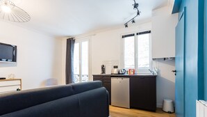 A compact living space with a sofa, kitchenette, and a TV mounted on the wall. - Cozy Studio in the Heart of Paris (Paris)