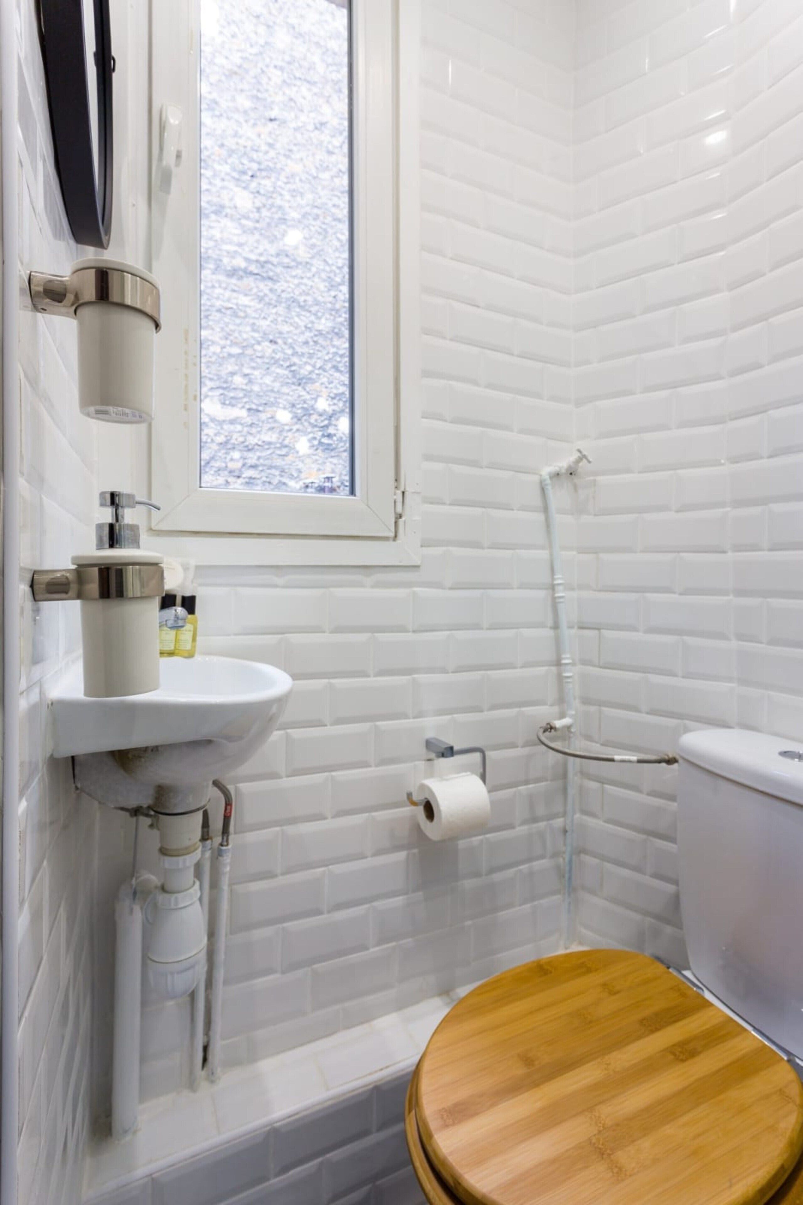 A bathroom with white tiled walls, a wooden toilet, a white sink with a faucet, and a window above the sink.