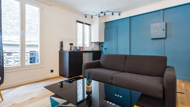 A modern living room with a sofa, a glass coffee table, and a kitchen area with cabinets and a window. - Cozy Studio in the Heart of Paris (Paris)