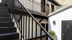 Eine Treppe führt zu einem Balkon mit Holzboden, einer schwarzen Tür und einer kleinen Pflanze.