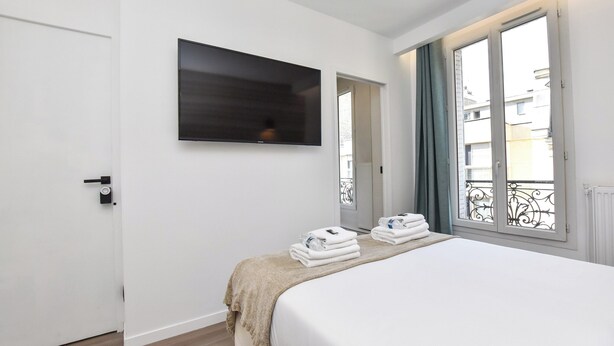 A hotel room with a bed, a flat-screen TV mounted on the wall, a window with a view of buildings, and a door. - Intimate Studio - 2P - Beaugrenelle/tour Eiffel (Paris)