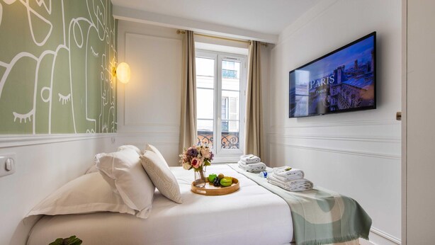 Une chambre d’hôtel avec un lit, une télévision, une fenêtre avec des rideaux, un mur orné d’un tableau et un plateau avec des serviettes et des fruits.