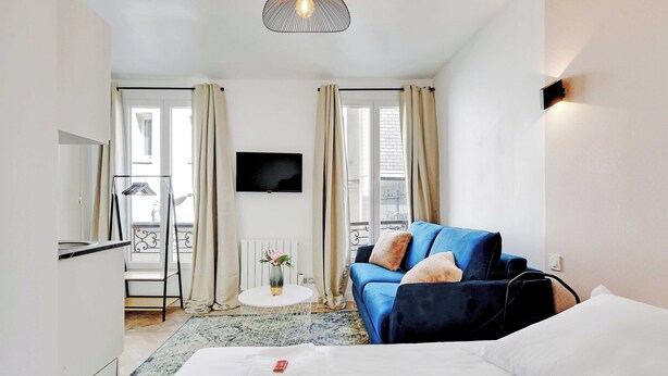Habitación de hotel con cama, un sofá azul, televisor, una mesita con un florero y balcón con barandilla.