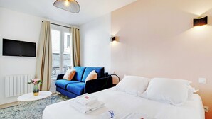 Una habitación de hotel con una cama, un sofá azul, una mesita redonda con un florero y un televisor montado en la pared.