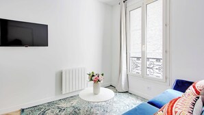 Una sala de estar moderna con un televisor de pantalla plana montado en la pared, una mesa de centro blanca y redonda con un jarrón de flores, un sofá azul con cojines estampados, y un gran ventanal con vistas al balcón.