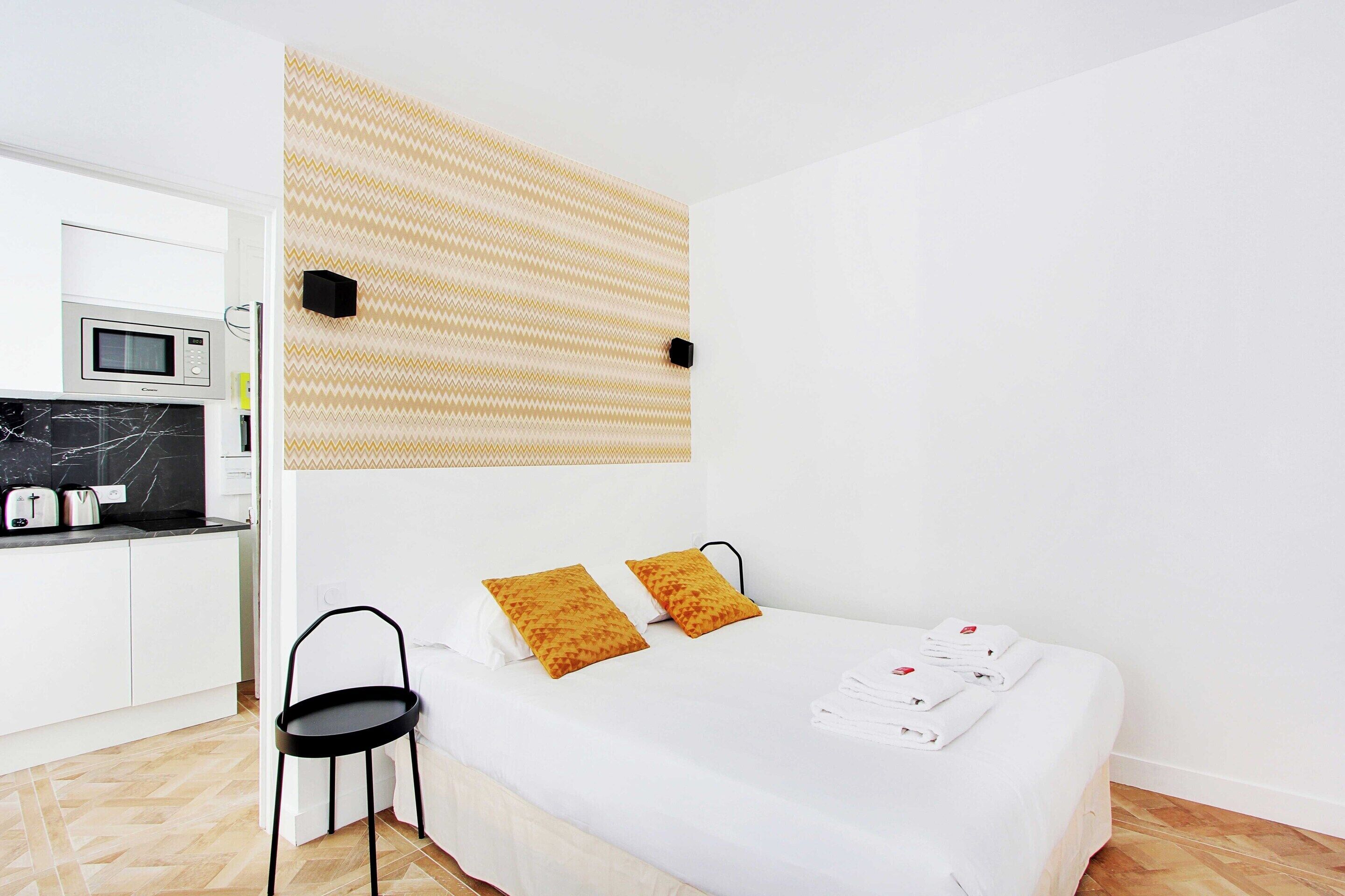 A modern hotel room with a bed, two pillows, a bedside table, and a microwave in the adjacent kitchenette.