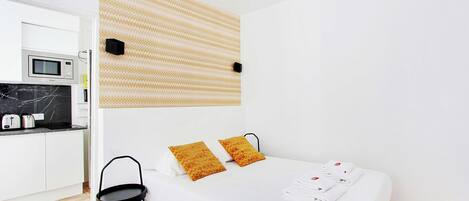 A modern hotel room with a bed, two pillows, a bedside table, and a microwave in the adjacent kitchenette.