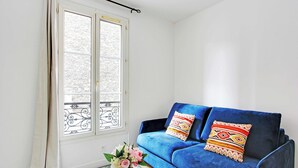 Una habitación con un sofá azul, un jarrón blanco lleno de flores y una ventana con cortinas.