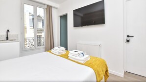 A hotel room with a bed, a TV mounted on the wall, a window with curtains, and a view of buildings outside.