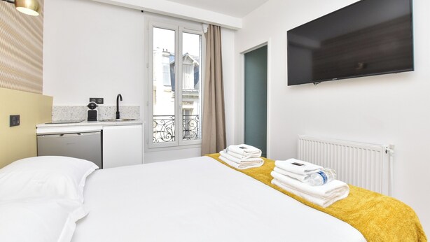 A hotel room with a bed, a TV mounted on the wall, a small kitchenette, and a window with a view of buildings.