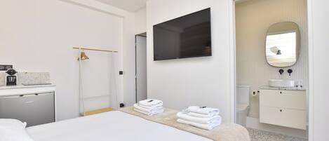A hotel room with a bed, a TV mounted on the wall, a bathroom with a sink and mirror, and a small desk with a coffee maker.