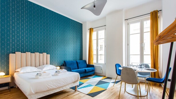 Habitació d'hotel moderna amb un llit, un sofà blau, una taula de menjador amb cadires i una finestra amb cortines.