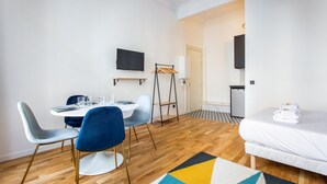 Habitació d'hotel moderna amb terra de fusta, un llit amb roba de llit blanca, zona de menjador amb taula rodona i cadires, i un televisor muntat a la paret.