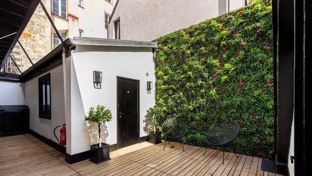 Ein Innenhof mit Holzdeck, einem weißen Gebäude, einer schwarzen Tür, einem Feuerlöscher, einem Blumentopf und einer mit Grünpflanzen und roten Blumen bewachsenen Mauer.