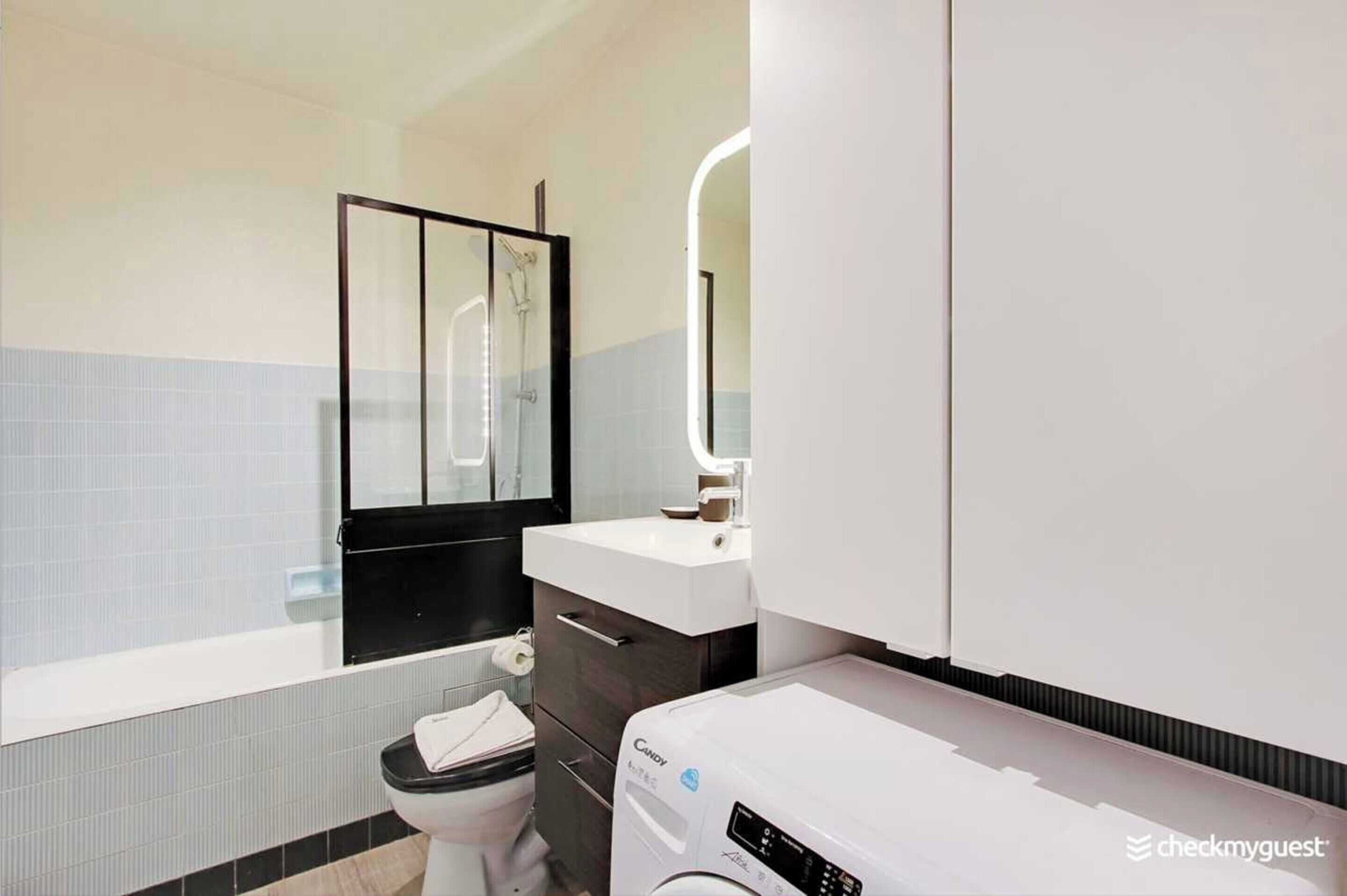 Uma casa de banho moderna com cabine de duche em vidro, lavatório branco, sanita e máquina de lavar roupa com abertura frontal.