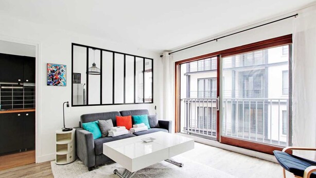 Un soggiorno moderno con un divano grigio, cuscini colorati, un tavolino da caffè bianco e un'ampia finestra vista edifici.