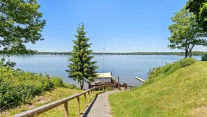 A wooden pathway leading to a dock by a calm lake, surrounded by greenery and a clear sky.