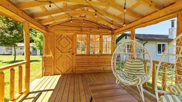 Une véranda en bois avec un toit abrité, un fauteuil suspendu et une vue sur une maison au loin.