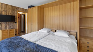A bedroom with a wooden headboard, a bed with white bedding, a TV mounted on the wall, and a wooden wardrobe. - Cosy studio with breathtaking view (Saint-Luc)