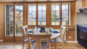 A dining area with a wooden table and chairs, large windows with a mountain view, and a kitchen area with a stove. - Cosy studio with breathtaking view (Saint-Luc)