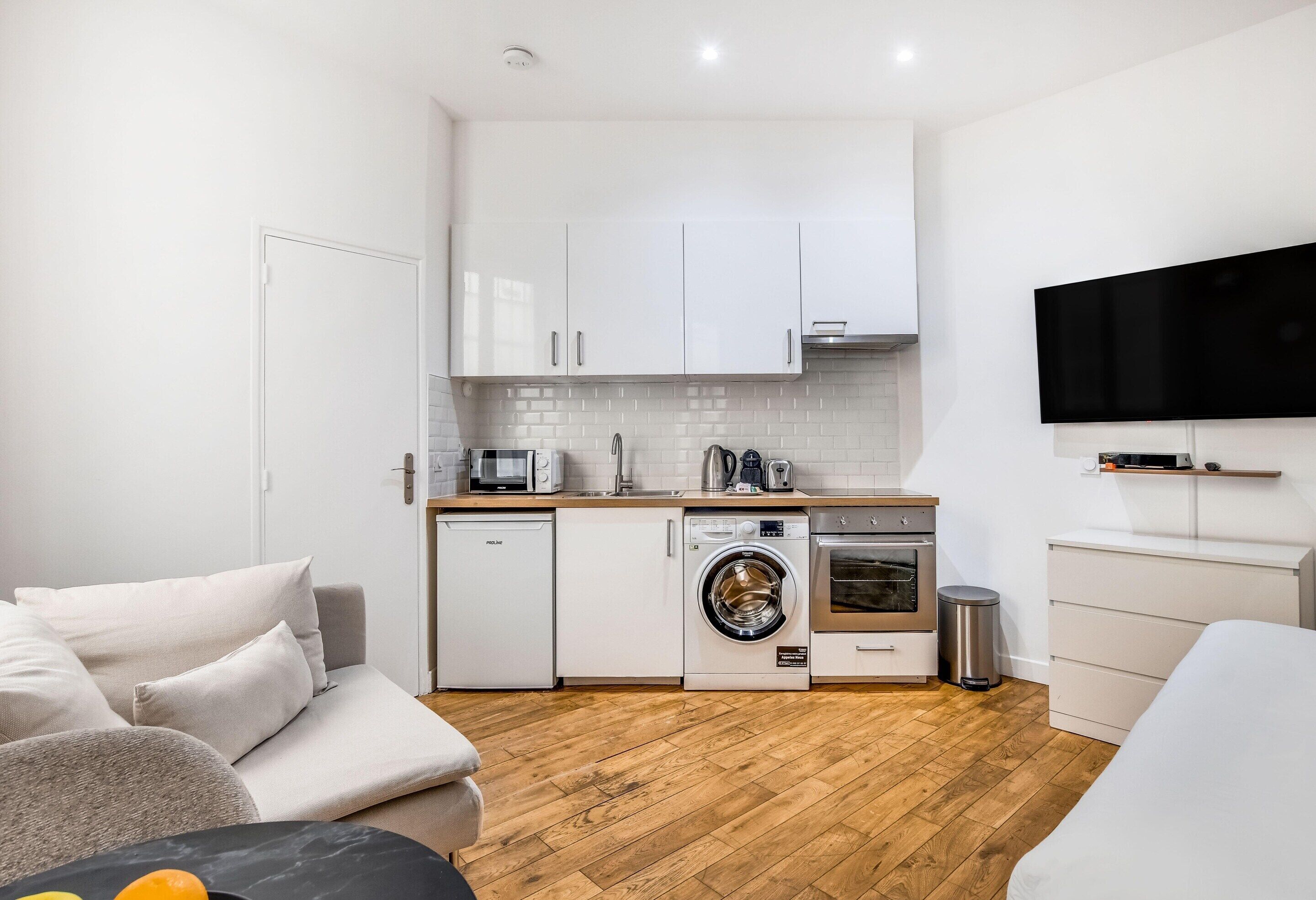 Cocina compacta con alacenas blancas, horno integrado, lavadora y televisor de pantalla plana montado en la pared.