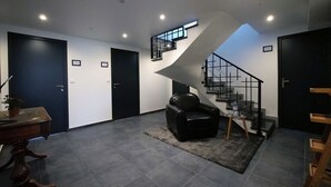 Un salon moderne avec un canapé en cuir noir, une petite table sur laquelle se trouvent une bouteille et un verre, et un escalier doté d’une rampe en métal.
