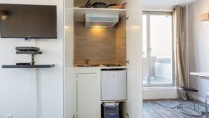 Ett kompakt hotellrum med en inbyggd TV, ett litet pentry, ett fönster med gardiner och en matplats.