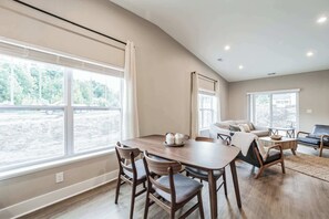 A spacious living room with a large dining table, comfortable seating, and large windows offering a view of the outdoors.